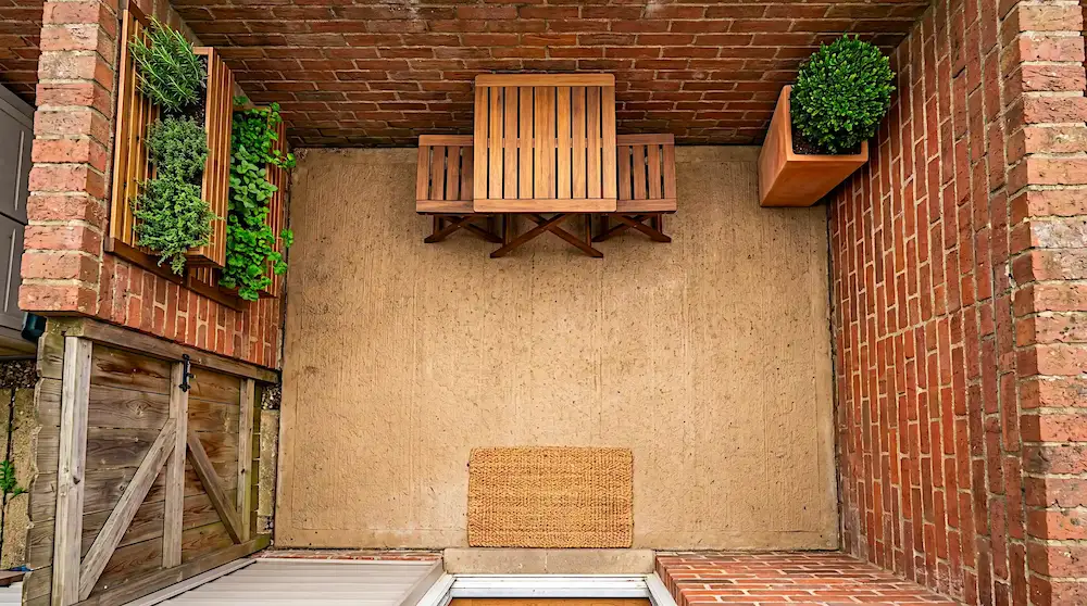 Overhead view of a tiny 6x8 foot brick patio with a small folding table, two folding stools tucked underneath, a terracotta topiary pot, and a natural jute doormat