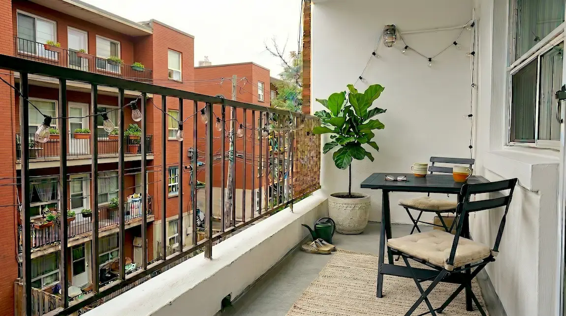 Small apartment balcony at dusk with warm Edison string lights clipped to a metal railing and a cream striped jute outdoor rug on the concrete floor