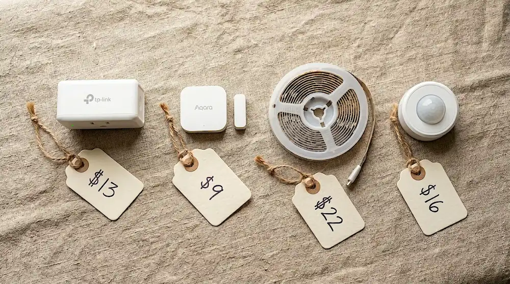A close-up overhead flat-lay of four budget smart home gadgets on a linen surface — a smart plug, a door sensor, a LED strip coiled, and a motion sensor, each with a handwritten price tag showing prices under $25