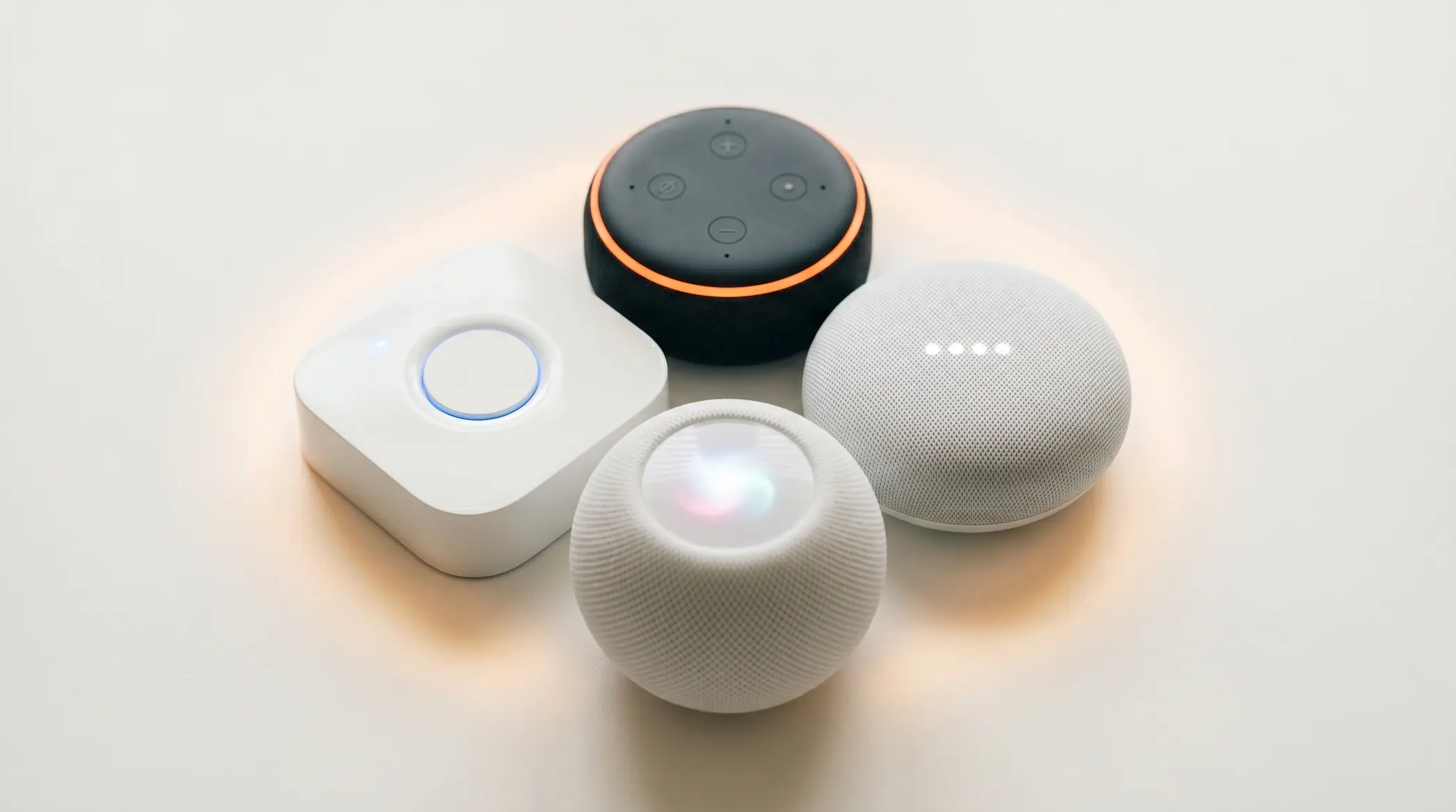 Four different smart home brand devices arranged in a circular cluster on a white surface — Amazon Echo Dot with orange ring, Philips Hue Bridge with blue ring, Apple HomePod mini with rainbow Siri glow, and Google Nest Mini with white dots all indicator lights glowing