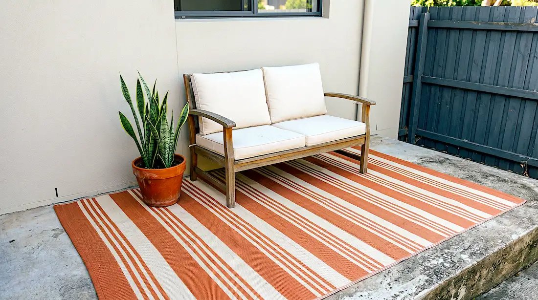 A flat-woven terracotta and cream striped outdoor rug on a small concrete patio with a wooden bench and snake plant in a terracotta pot, raw concrete visible at one edge