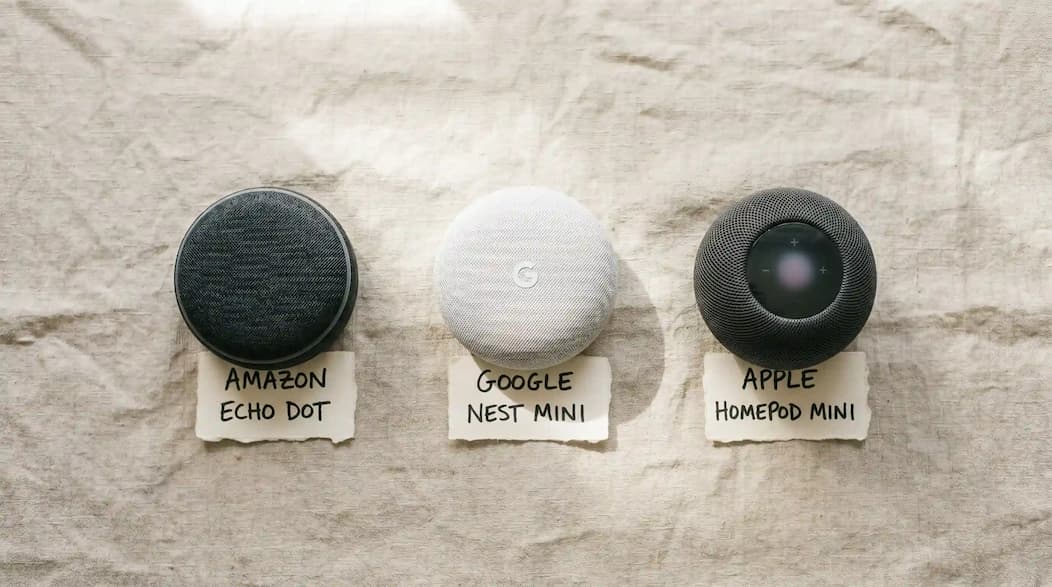 Amazon Echo Dot, Google Nest Mini, and Apple HomePod mini placed side by side on a concrete surface for comparison